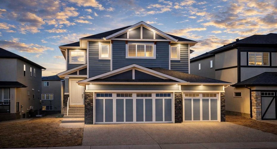 An architecturally striking Kinniburgh residence illuminated by premium EXTERIOR FEATURE LIGHTING, highlighting the LUXURY STONE AND WOOD EXTERIOR, expansive 6-CAR DRIVEWAY, and OVERSIZED TRIPLE ATTACHED GARAGE with dramatic evening presence.