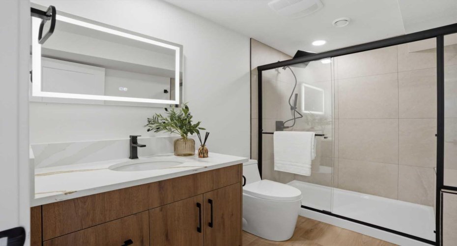 Stylish washroom with a fully tiled tub/shower combo, quartz vanity, backlit mirror, and upgraded black hardware for a clean, contemporary finish.