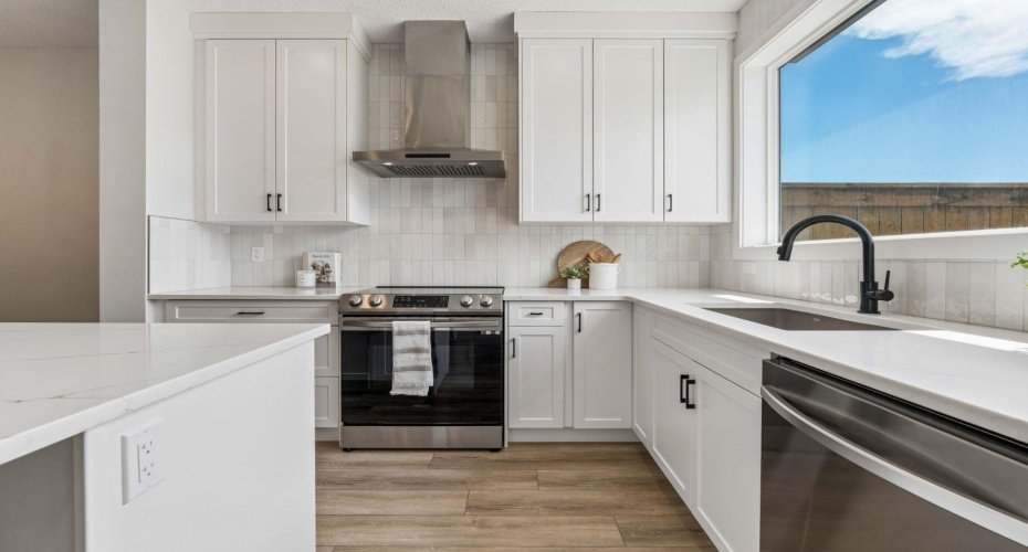 Designed with both style and function in mind, the kitchen showcases white shaker cabinetry, quartz countertops, a modern tile backsplash, stainless steel appliances, and a striking black farmhouse sink overlooking the backyard.