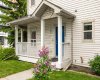 A welcoming covered porch with white columns and a bold blue door, surrounded by blooming lilac and tidy landscaping just steps from the shops of Marda Loop.
