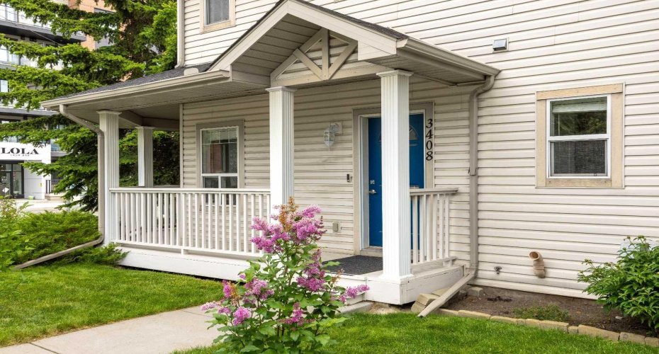 A welcoming covered porch with white columns and a bold blue door, surrounded by blooming lilac and tidy landscaping just steps from the shops of Marda Loop.