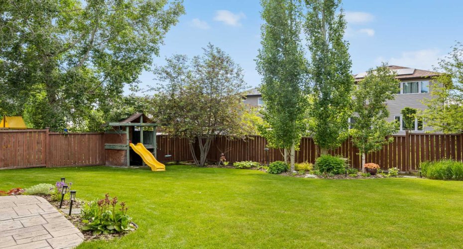 A kids' playhouse tucked to the side, mature trees for shade, and garden beds all around — this corner of the yard was made for childhood.