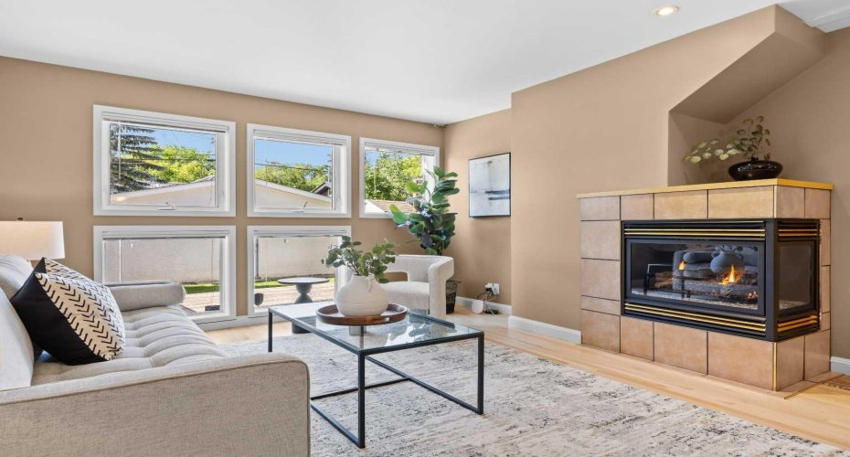 Tall windows fill the living room with sunlight, framing the fireplace as the room's natural focal point.